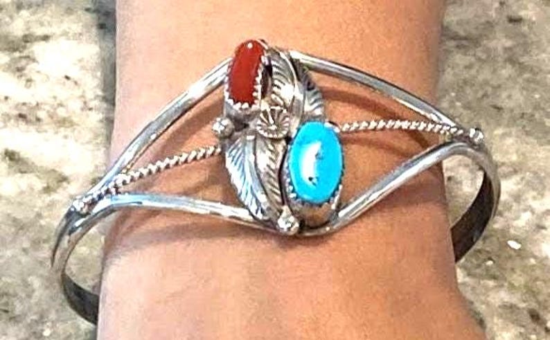 May include: Silver cuff bracelet with a central design featuring a red stone, a turquoise stone, and silver feather accents. The bracelet has a split band design and is worn on a wrist.