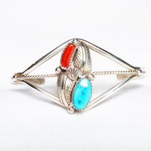May include: Silver cuff bracelet with a central design featuring a red coral and a turquoise stone, flanked by detailed silver leaf accents. The bracelet has a geometric open design with a twisted rope detail.