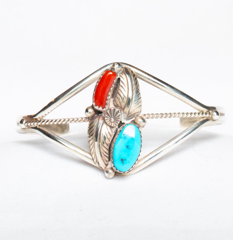 May include: Silver cuff bracelet with a central design featuring a red coral and a turquoise stone, flanked by detailed silver leaf accents. The bracelet has a geometric open design with a twisted rope detail.
