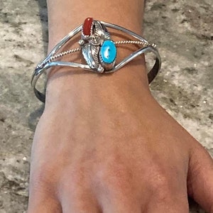 May include: A silver cuff bracelet with a central design featuring a red stone and a turquoise stone. The bracelet has a detailed, ornate design with a feather motif. A silver ring with a decorative pattern is also visible.