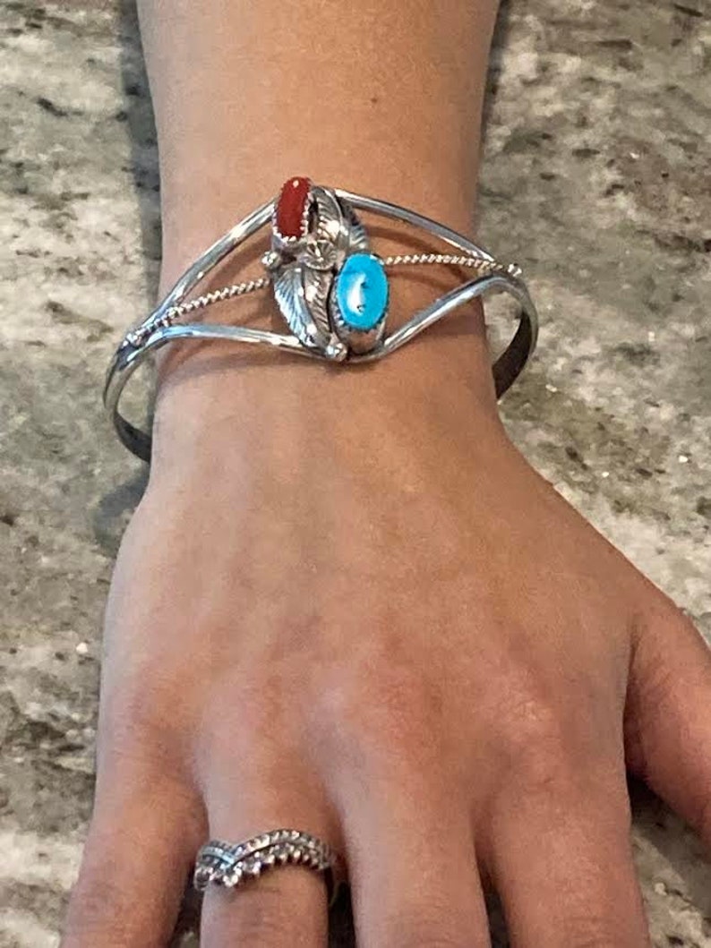 May include: A silver cuff bracelet with a central design featuring a red stone and a turquoise stone. The bracelet has a detailed, ornate design with a feather motif. A silver ring with a decorative pattern is also visible.