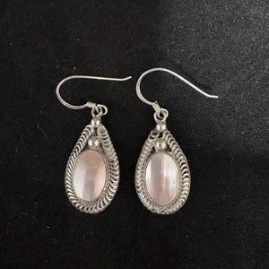 May include: A pair of silver-toned drop earrings. Each earring features a pearlescent, oval-shaped center stone, framed by a detailed, textured border. The earrings have a hook closure.