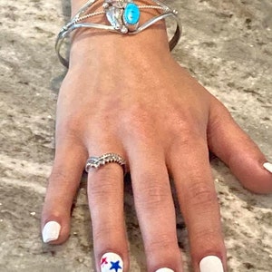 May include: A silver cuff bracelet with turquoise and coral accents, and a ring with a decorative design. The nails are painted white, with a star design on one nail.
