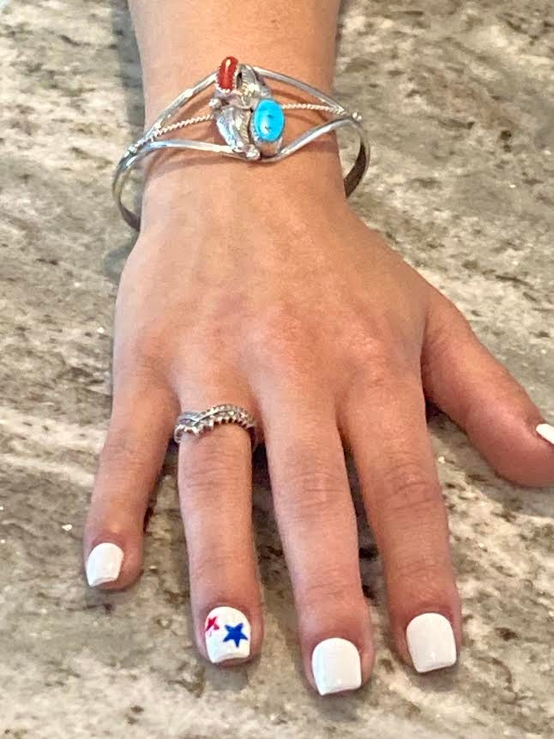 May include: A silver cuff bracelet with turquoise and coral accents, and a ring with a decorative design. The nails are painted white, with a star design on one nail.