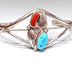 May include: Silver cuff bracelet with a central design featuring a red coral and a turquoise stone. The bracelet has a diamond-shaped frame with a twisted rope detail and feather accents, showcasing a Native American style.