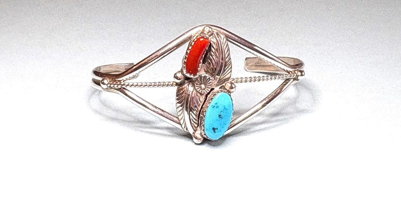 May include: Silver cuff bracelet with a central design featuring a red coral and a turquoise stone. The bracelet has a diamond-shaped frame with a twisted rope detail and feather accents, showcasing a Native American style.