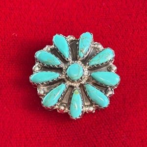 May include: A silver-toned flower-shaped brooch with turquoise stone petals and a central turquoise stone. The brooch is set against a red background, showcasing the vibrant colors and intricate design.