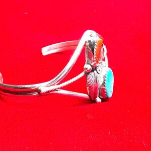 May include: Silver cuff bracelet with a turquoise and orange stone setting. The bracelet features a detailed design with a feather and floral accents. The background is a solid red.