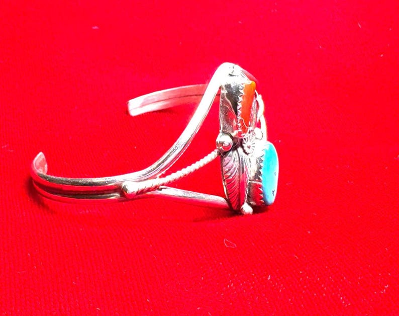 May include: Silver cuff bracelet with a turquoise and orange stone setting. The bracelet features a detailed design with a feather and floral accents. The background is a solid red.