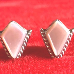 May include: A pair of diamond-shaped stud earrings. Each earring features a light pink, pearlescent center stone set in a silver-toned metal frame with a twisted rope design around the edges. The earrings are displayed on a red surface.