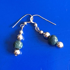 May include: A pair of drop earrings featuring silver-toned metal hooks and a vertical arrangement of alternating polished silver-toned spherical beads and textured, dark green, rounded beads. The design includes three silver beads and two green beads.