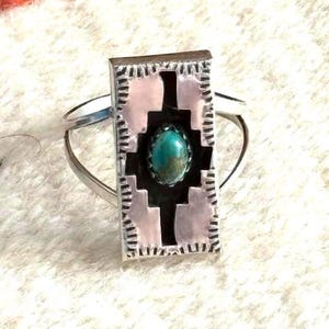 May include: A silver ring with a rectangular turquoise stone set in a decorative frame. The frame features a geometric design with pink and black accents, creating a Southwestern aesthetic. The ring's band is split on either side of the frame.