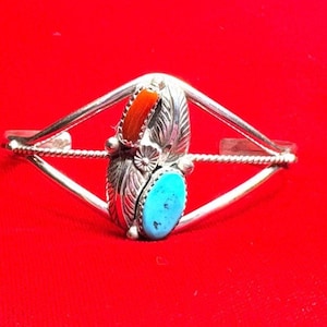 May include: Silver-toned cuff bracelet with a central design featuring a turquoise oval stone and a coral-colored rectangular stone. The bracelet has a leaf-shaped element and a floral accent.