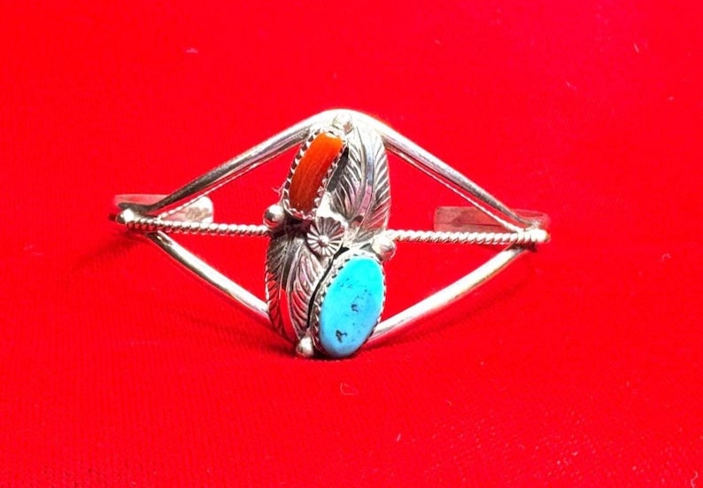 May include: Silver-toned cuff bracelet with a central design featuring a turquoise oval stone and a coral-colored rectangular stone. The bracelet has a leaf-shaped element and a floral accent.