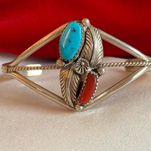 May include: Silver cuff bracelet with turquoise and red coral stones. The bracelet features a central design with a turquoise oval stone at the top and a red coral stone at the bottom, framed by detailed silver leaves.