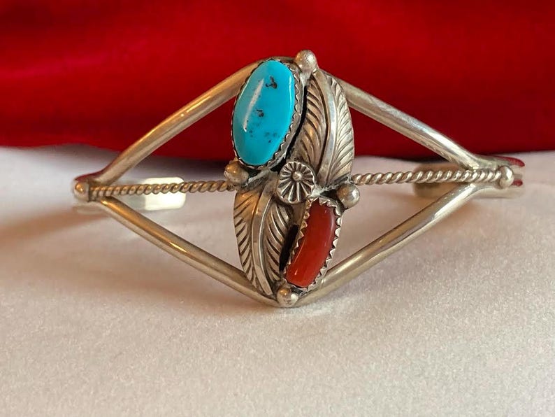 May include: Silver cuff bracelet with turquoise and red coral stones. The bracelet features a central design with a turquoise oval stone at the top and a red coral stone at the bottom, framed by detailed silver leaves.