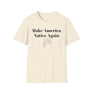 May include: Cream-colored t-shirt with the text "Make America Native Again" in a bold, serif font. Below the text is a black illustration of wheat stalks. The shirt is made of a soft, textured fabric.