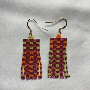 Może przedstawiać: Para kolczyków z koralików z geometrycznym wzorem w odcieniach różowego, pomarańczowego i żółtego. Kolczyki mają frędzle i złote haczyki.