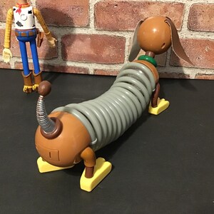 May include: A Slinky Dog toy from the Toy Story franchise. The toy dog has a brown head and body with a gray slinky-like midsection. It has yellow feet and a green collar. A Woody action figure is in the background.