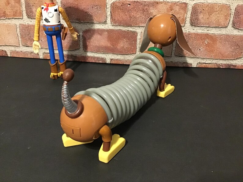 May include: A Slinky Dog toy from the Toy Story franchise. The toy dog has a brown head and body with a gray slinky-like midsection. It has yellow feet and a green collar. A Woody action figure is in the background.