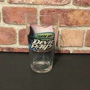 May include: A clear glass with a Tampa Bay Devil Rays logo. The logo is blue, green, and black with the words "Tampa Bay Devil Rays".