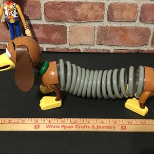May include: A Slinky Dog toy with a brown head, ears, and body, a gray spring midsection, and yellow feet. A Woody action figure stands behind the dog. A wooden ruler is in the foreground.