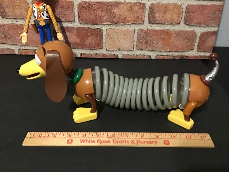May include: A Slinky Dog toy with a brown head, ears, and body, a gray spring midsection, and yellow feet. A Woody action figure stands behind the dog. A wooden ruler is in the foreground.