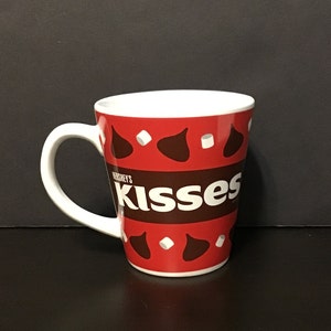 May include: A red and white ceramic mug with a Hershey's Kisses design. The mug features a brown and white pattern of Hershey's Kisses and white marshmallows. The text "Hershey's Kisses" is printed in white on a brown stripe.