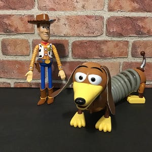 May include: Two Toy Story character figures: Sheriff Woody in a cowboy hat and Slinky Dog, a dachshund with a spring body. Woody wears a yellow shirt, vest, blue pants, and brown boots. Slinky Dog is brown, yellow, and gray.