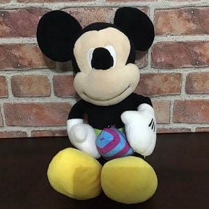 May include: A plush Mickey Mouse toy sitting with a colorful ball in its hands. The toy is black, yellow, and beige with black eyes and a big smile.