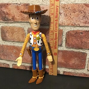 May include: A Woody action figure, featuring a brown cowboy hat, yellow plaid shirt, and blue pants. The figure has a sheriff's badge and a holster with a toy gun. The figure is approximately 8 inches tall.