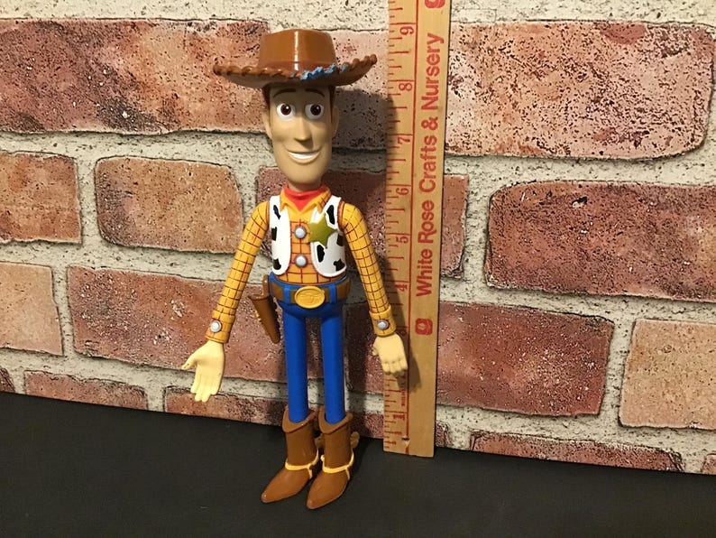 May include: A Woody action figure, featuring a brown cowboy hat, yellow plaid shirt, and blue pants. The figure has a sheriff's badge and a holster with a toy gun. The figure is approximately 8 inches tall.
