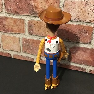 May include: A Woody action figure, featuring a brown cowboy hat, yellow plaid shirt, white vest with black spots, blue pants, and brown boots. The figure has a red neckerchief and a brown holster.