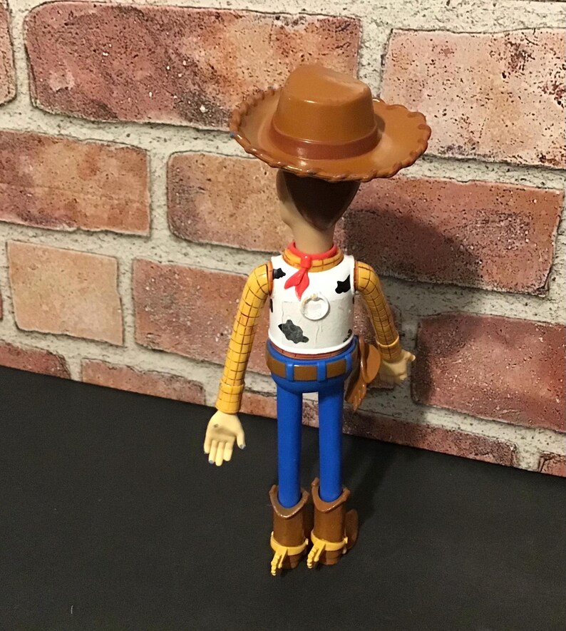 May include: A Woody action figure, featuring a brown cowboy hat, yellow plaid shirt, white vest with black spots, blue pants, and brown boots. The figure has a red neckerchief and a brown holster.