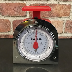 May include: A silver and red kitchen scale with a white dial that reads "TAYLOR" and has markings for pounds and kilograms. The scale has a capacity of 6 lbs / 2.7 kg.