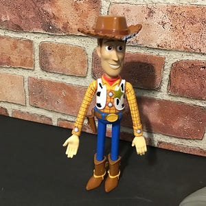 May include: A Woody action figure, featuring a brown cowboy hat, yellow plaid shirt, blue pants, and brown boots. The figure has a sheriff's badge and a holster. The toy is standing in front of a brick wall.