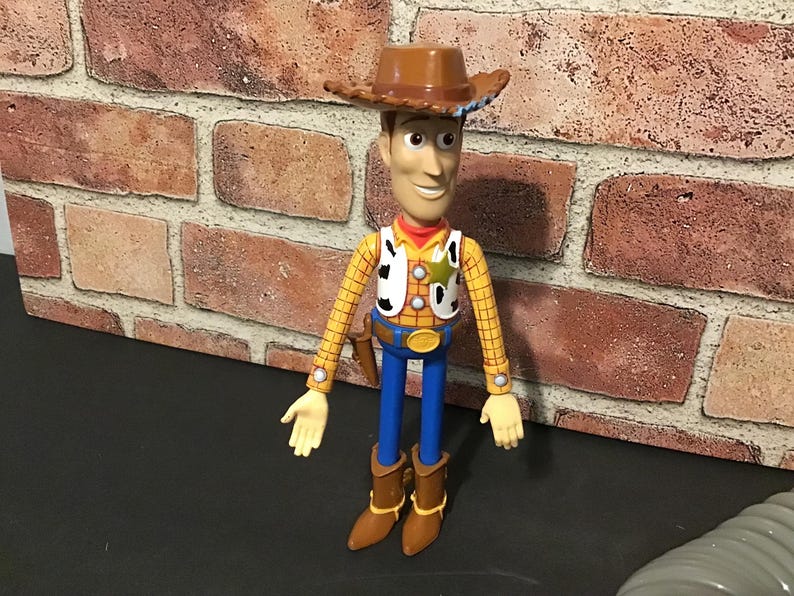 May include: A Woody action figure, featuring a brown cowboy hat, yellow plaid shirt, blue pants, and brown boots. The figure has a sheriff's badge and a holster. The toy is standing in front of a brick wall.