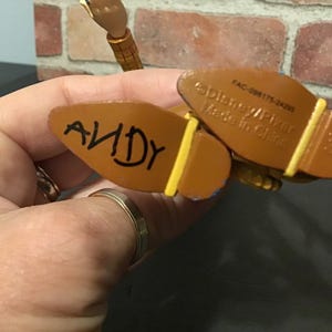 May include: Close-up of the bottom of a Woody action figure's boots. The word "ANDY" is written in black marker on the sole of one boot. The other boot has text that reads "Disney/Pixar Made in China".