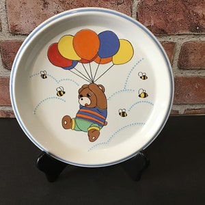 May include: A white ceramic plate with a blue rim features a cartoon bear holding onto colorful balloons. The bear is wearing a blue and orange striped shirt and green pants. The plate is decorated with small yellow bees and blue dots.