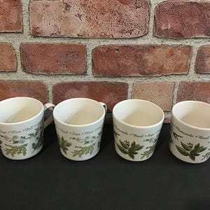 May include: Four white ceramic mugs with green and brown herbal designs. The mugs have text that reads "Mint, Dill, Sage, Basil, Chamomile".
