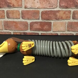 May include: A Slinky Dog toy from the Toy Story franchise. The toy dog has a brown head and body with yellow ears and feet. The body is a gray slinky-like coil. The toy is lying on a black surface.