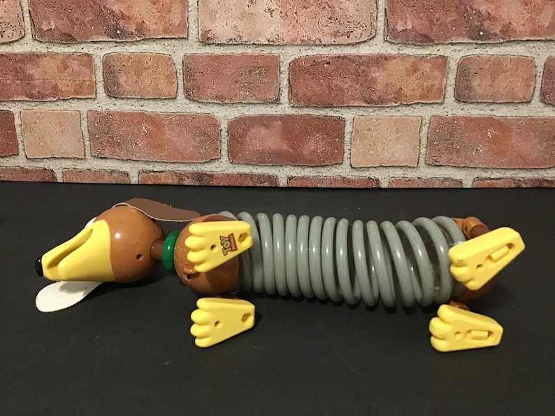 May include: A Slinky Dog toy from the Toy Story franchise. The toy dog has a brown head and body with yellow ears and feet. The body is a gray slinky-like coil. The toy is lying on a black surface.