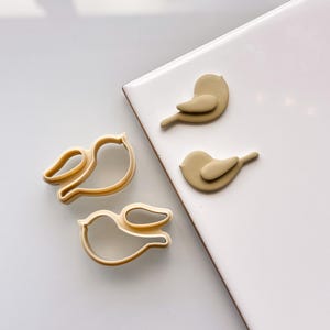 May include: Two cookie cutters and two fondant birds in the shape of birds. The cookie cutters and fondant birds are beige.