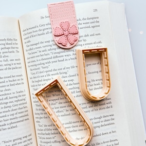 以下が含まれることがあります： ピンク色の革製ブックマーク、花の模様とベージュ色のU字型クッキーカッター2つ。すべて本の上に置かれています。ブックマークは本に挟まれています。