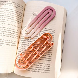 May include: Two large, colorful paperclip-shaped bookmarks rest on an open book. One bookmark is a gradient of pink and white, while the other is a solid peach color. The bookmarks are made of a smooth, possibly plastic material.