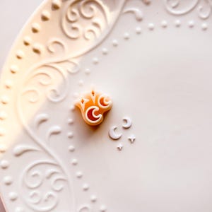May include: A white, ornate ceramic plate with a raised floral design. Small, orange-toned decorative elements in the shape of a crown, crescent moons, and stars are arranged on the plate.