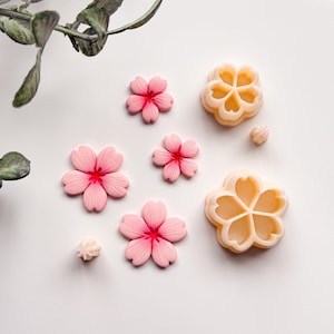 May include: A collection of pink and red flower-shaped decorations and molds. The decorations are in various sizes, with a light pink hue and red centers. Two beige molds are also present, along with a sprig of green leaves.