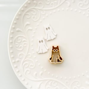 May include: Three Halloween-themed cookie cutters on a white plate. Two are white ghost shapes, and the third is a brown and tan cat shape. The plate has a decorative, embossed floral pattern.