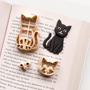 May include: An assortment of cat-themed items, including two tan cat-shaped cookie cutters, a black cat figurine, and a small tan butterfly-shaped item. The items are arranged on an open book.