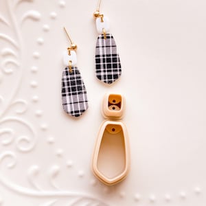 May include: A pair of dangle earrings with a black and white plaid pattern, white square accents, and gold-colored posts. A beige earring-shaped cutter is also present.